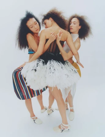 Three women in fashionable dresses pose in a bright studio. One model wears a tutu dress, and another a striped skirt with a cropped top.