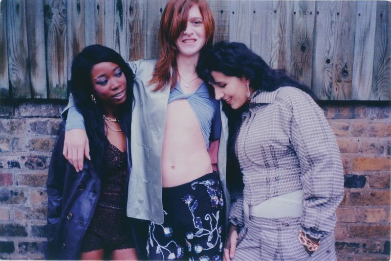 Three young women pose together outdoors in casual fashion, standing in front of a brick and wooden fence. Candid, lifestyle shot.