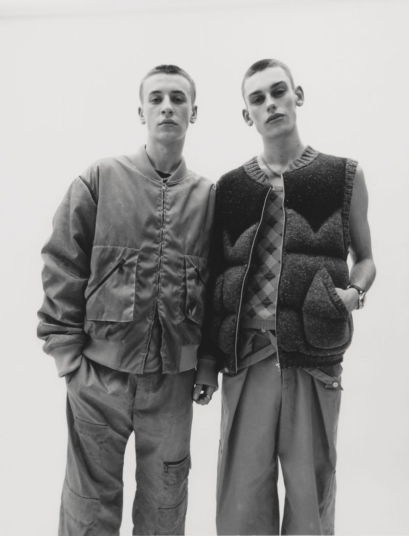 Two young adult males stand indoors against a white background, wearing casual fashion: a bomber jacket and a puffer vest.