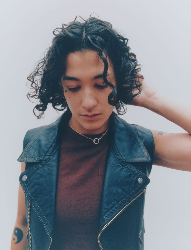 Indoor studio portrait of one adult with curly hair wearing a vest and necklace looking downward against a clean white background.