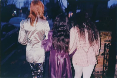 Three models with long dark hair walking away down an urban street at dusk, viewed from behind wearing coats.