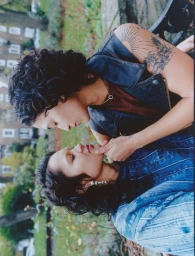 Two women sharing an intimate moment outdoors, one gently holding the other's chin while wearing denim and a dark vest.