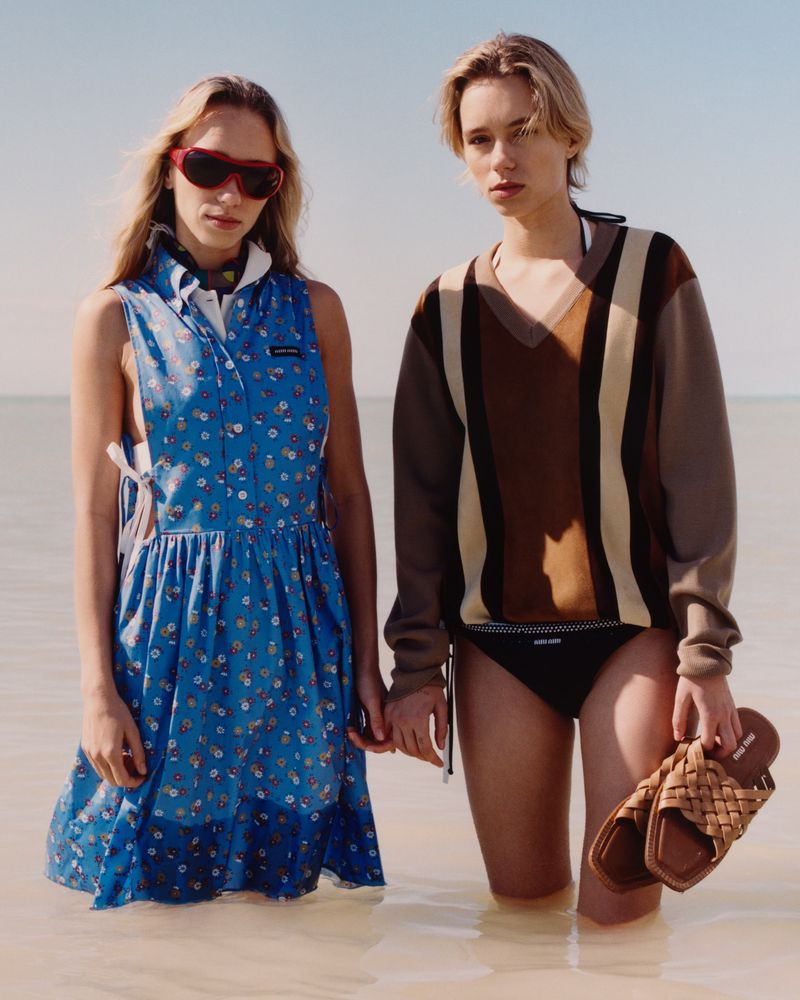 Two women wade in shallow water, wearing a dress and sweater, holding hands and sandals in a serene, sunlit beach scene.