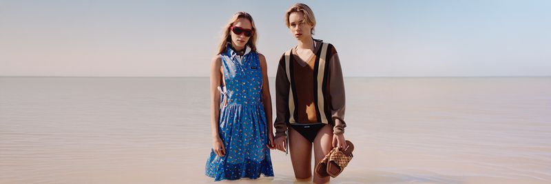Two women stand in shallow water, one in a blue floral dress and red sunglasses, the other in a striped sweater holding sandals.