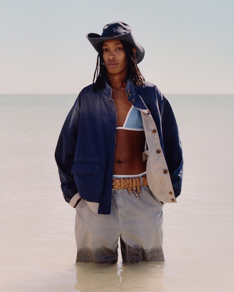 Person in water wearing a hat, bikini top, shorts, and jacket poses against a blurred horizon with blue sky for a fashion editorial.