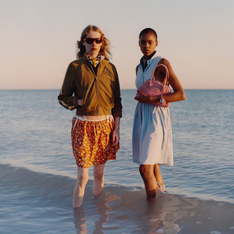 Two women stand in shallow ocean water wearing fashionable summer clothing and accessories, posing against the soft horizon.