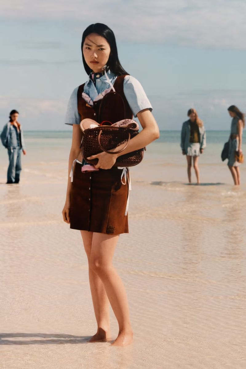 Fashion shot of a young Asian woman on a beach wearing a brown dress and holding a brown purse with people in background.
