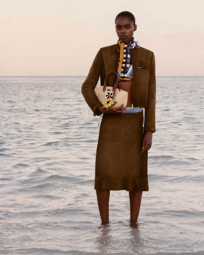 A woman in a brown jacket and skirt stands in ocean waves, holding a straw purse with a flower, against a background of sea and sky.