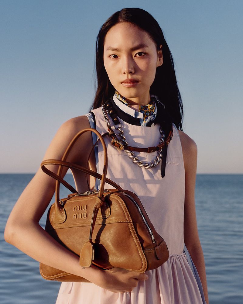 An Asian model poses outdoors against a blue sky and water, holding a brown leather Miu Miu handbag and wearing a light pink dress.