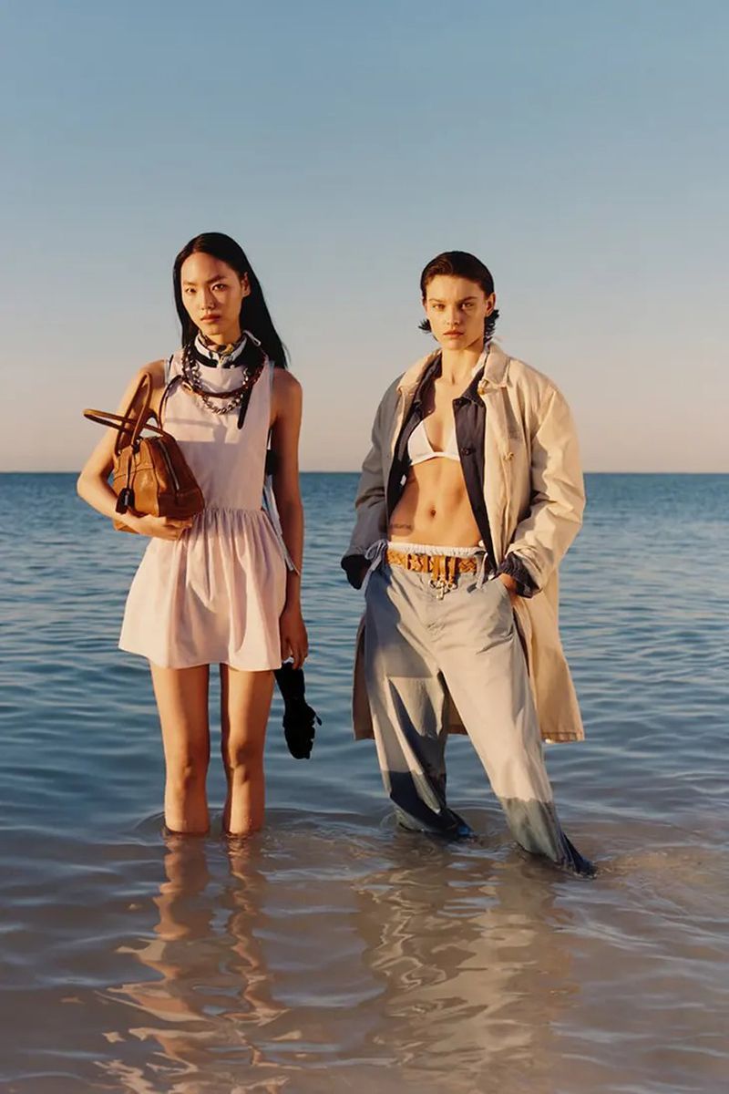 Two women in fashionable summer outfits stand in shallow ocean water, one holding a tan purse, under a clear blue sky on a sunny day.