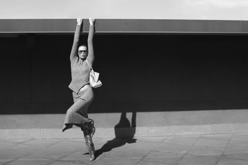 A woman in a monochrome outfit and boots hangs from a ledge in a black and white fashion shot with strong contrast and shadow.
