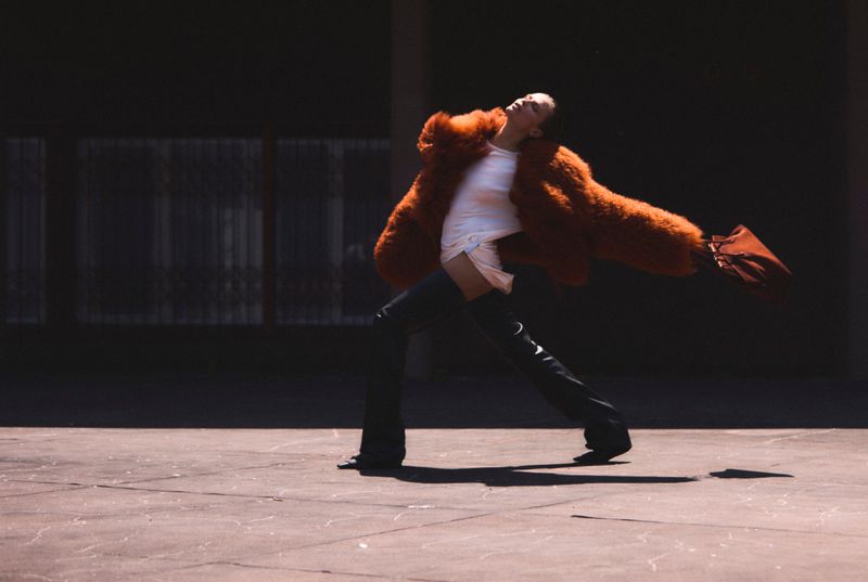 Woman in an orange fur coat and black thigh-high boots poses in an urban setting with concrete floors and a dark building backdrop.