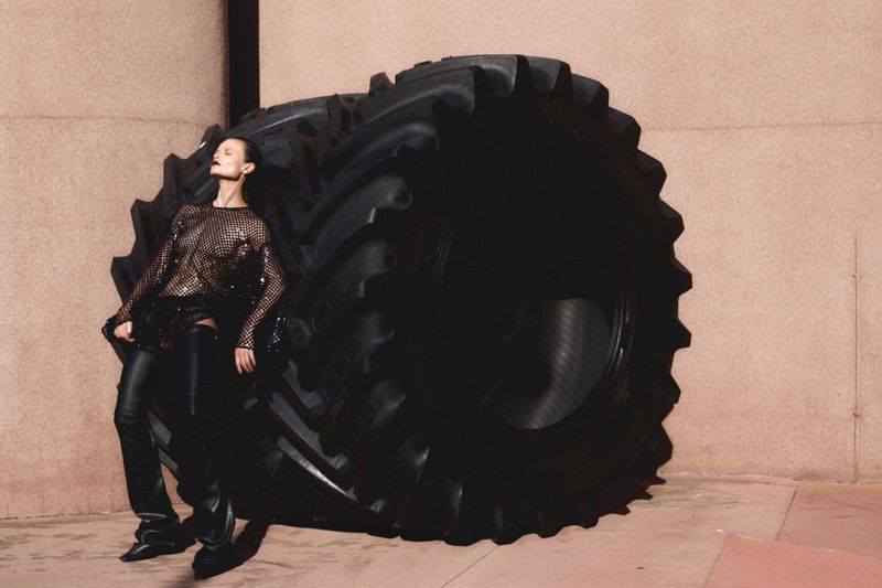 Woman posing in shiny top and black leather pants alongside a large tractor tire in an urban setting with neutral background.