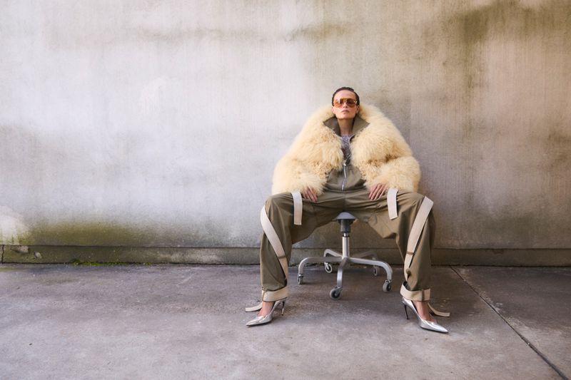 Fashionable adult female in sunglasses, fur coat, and heels poses while sitting on a rolling stool in front of a textured wall.