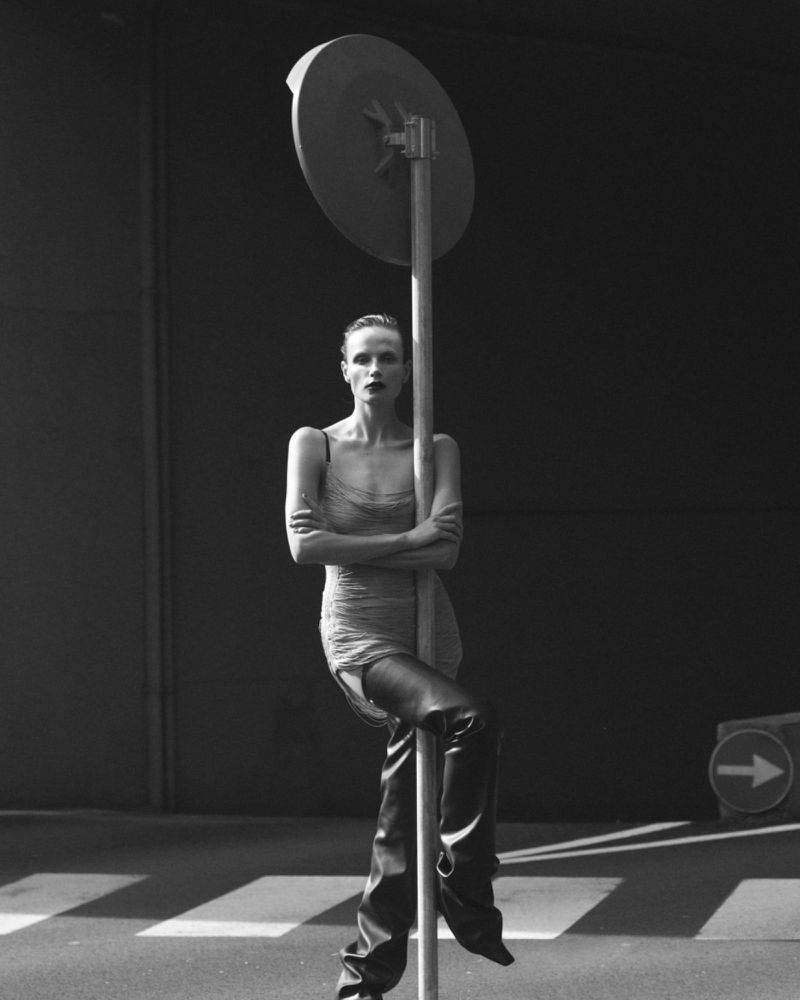 Monochrome fashion shot of a woman leaning on a signpost in a dress and over-the-knee boots on a crosswalk with a dark background.