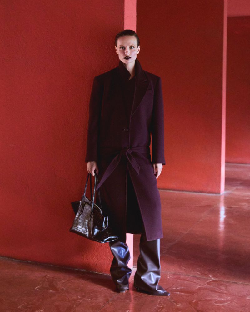 Fashionable woman in a plum coat stands against a red wall, holding a black alligator bag, showcasing modern style in an indoor setting.