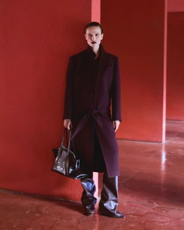 Fashionable woman in a plum coat stands against a red wall, holding a black alligator bag, showcasing modern style in an indoor setting.