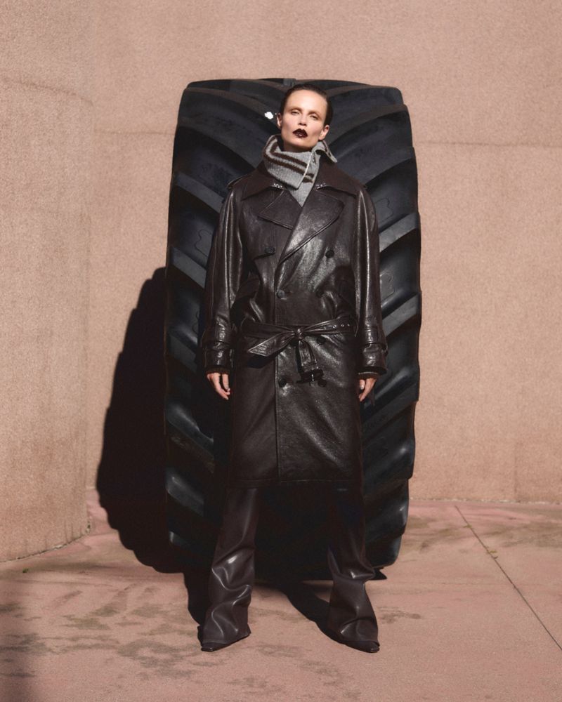 Fashion shot of an adult female wearing a dark brown leather trench coat, leather pants and standing next to a very large tire.