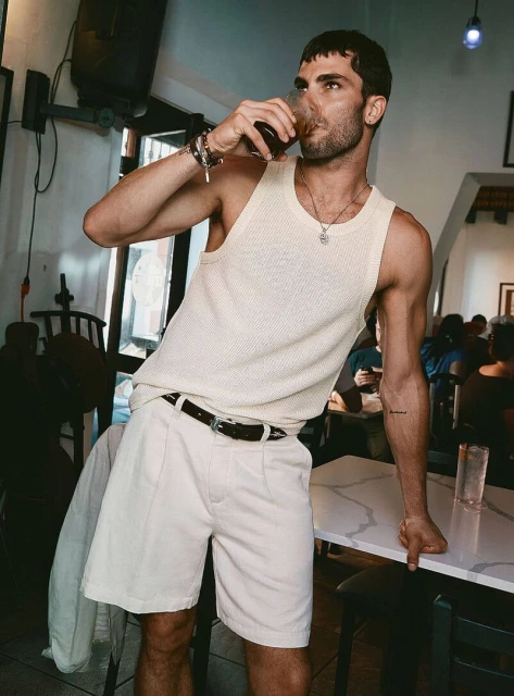 A young adult male drinks from a glass while leaning on a table in an indoor restaurant setting wearing a tank top and shorts.