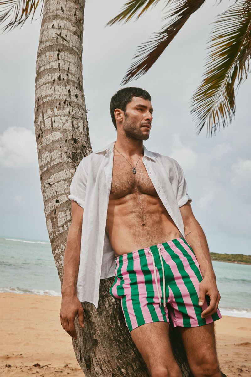 Male model wearing unbuttoned white shirt and green-and-pink striped swim trunks leaning against a palm tree on a sandy beach.