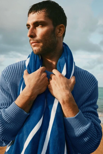 Man in blue sweater on beach, holding blue and white striped towel. Ocean and sky are visible in the background on a sunny day.