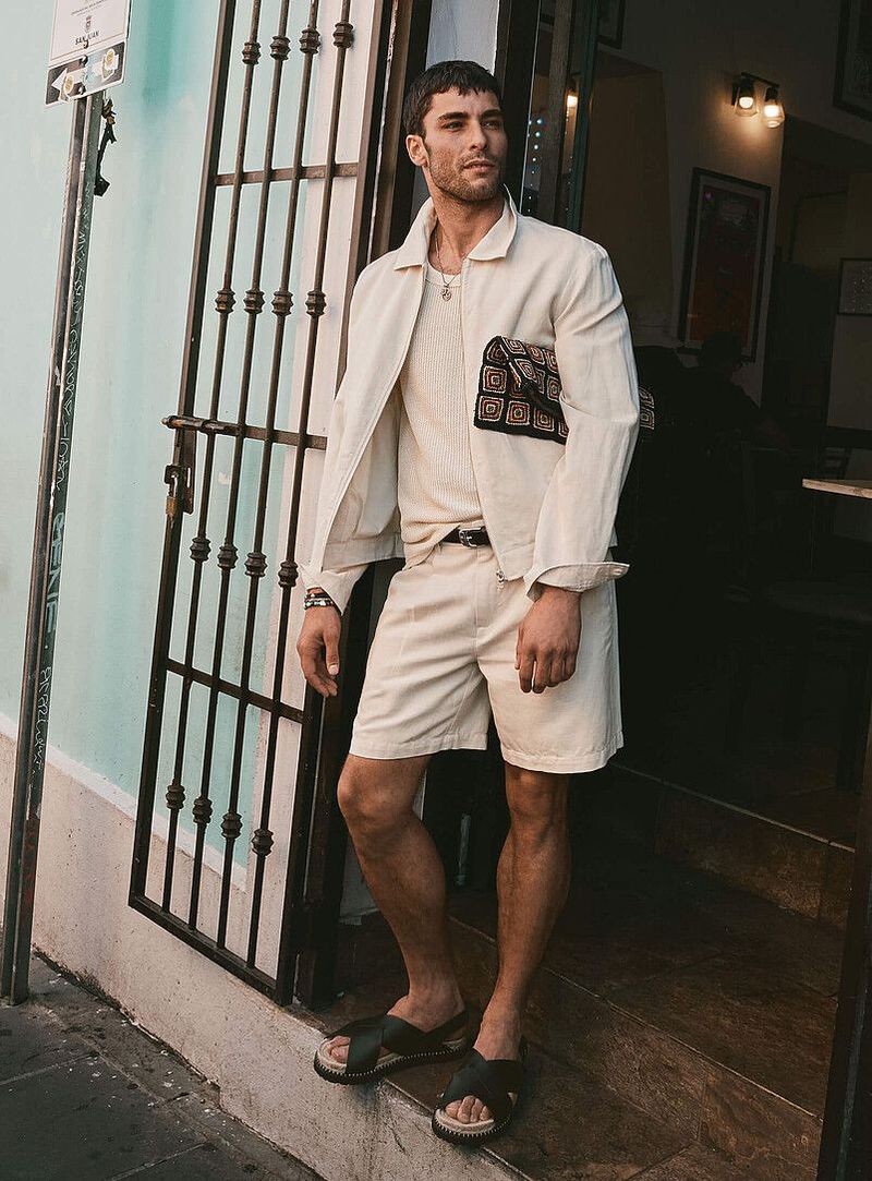 Model Paolo Busti in cream jacket and shorts holding patterned clutch outside doorway for Simons Canada