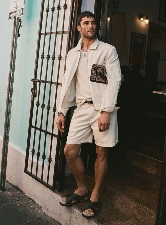 Model Paolo Busti in cream jacket and shorts holding patterned clutch outside doorway for Simons Canada