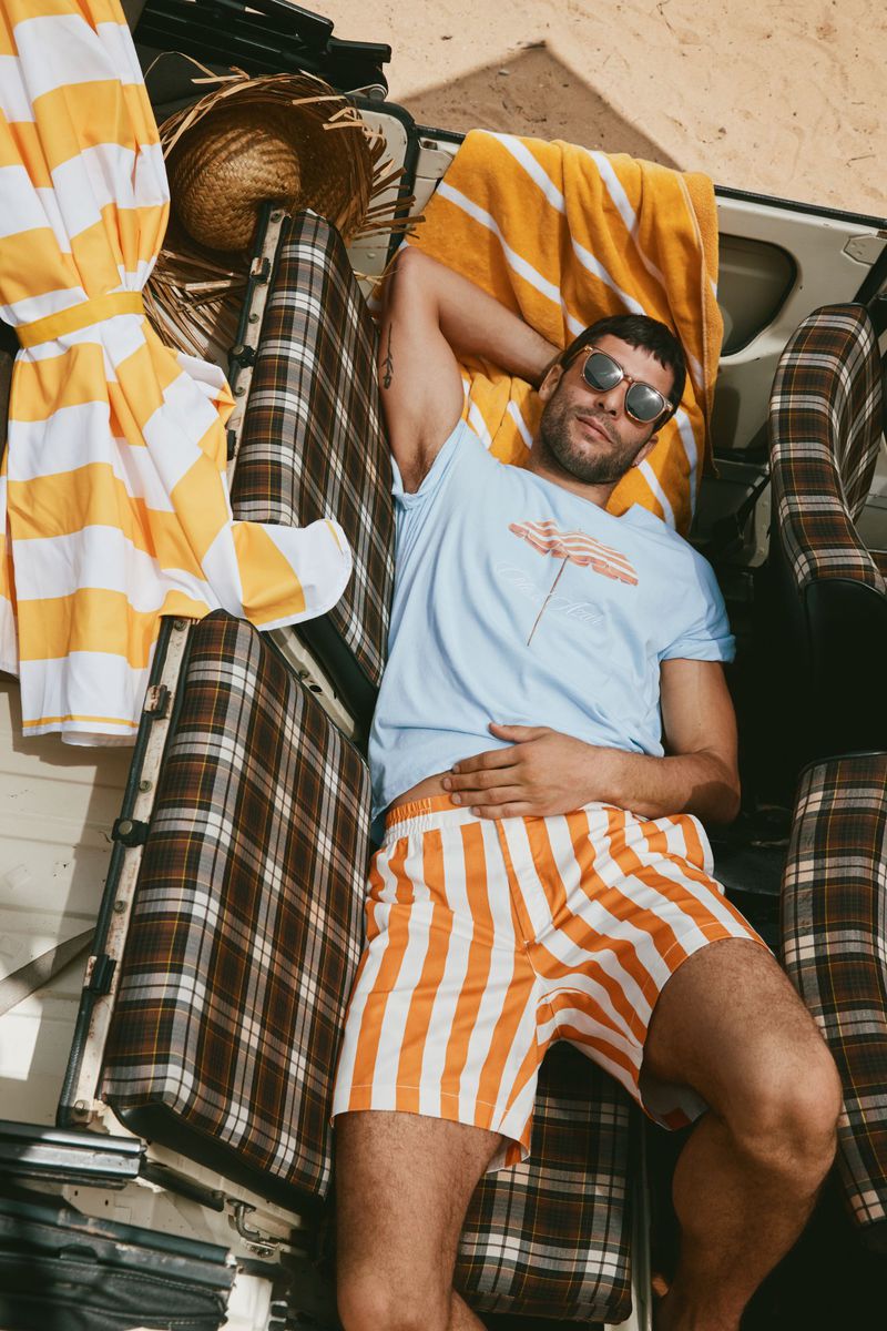 Adult man wearing sunglasses, striped shorts and t-shirt, lies on a van seat with yellow striped beach towels and a straw hat.