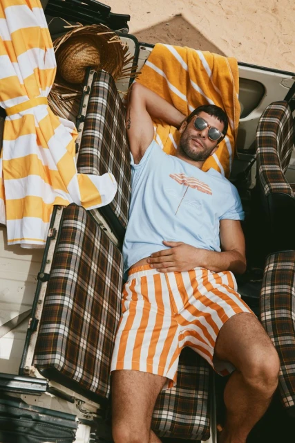 Adult man wearing sunglasses, striped shorts and t-shirt, lies on a van seat with yellow striped beach towels and a straw hat.