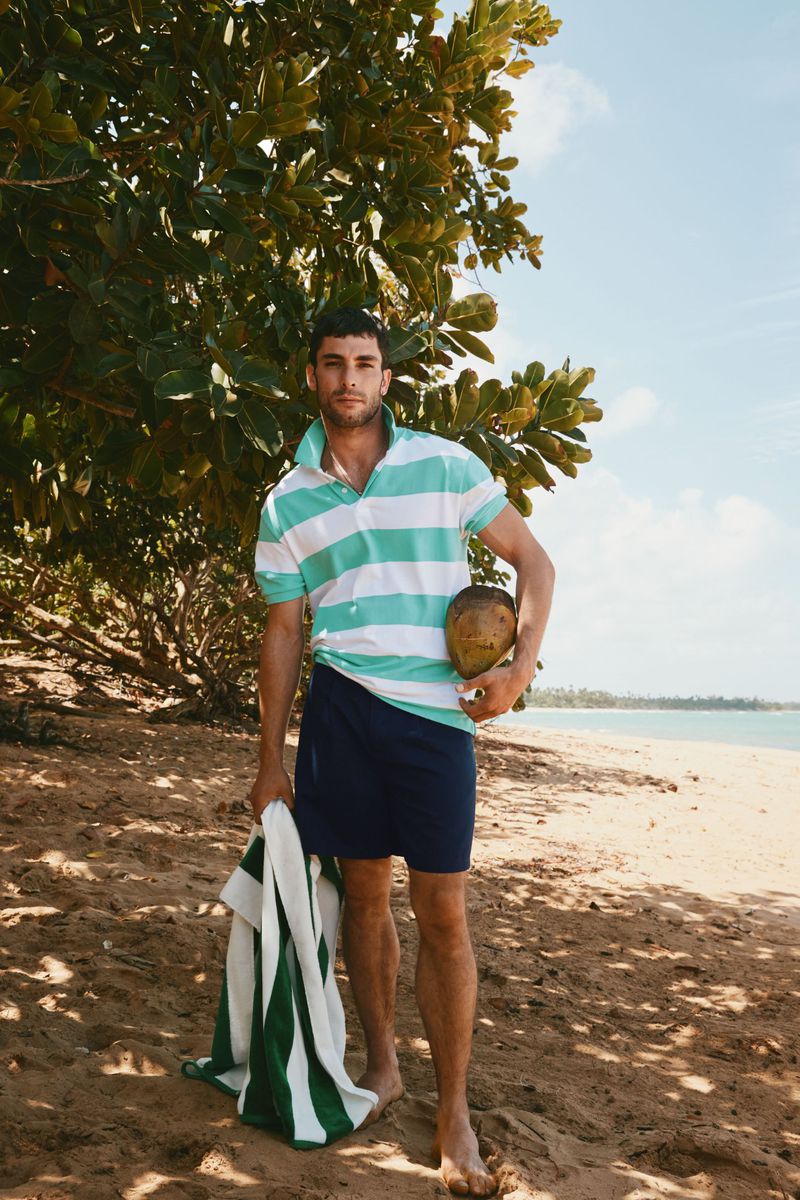 A tanned man poses on a sandy beach holding a coconut and striped towel, wearing a striped polo shirt, in sunny, outdoor setting.