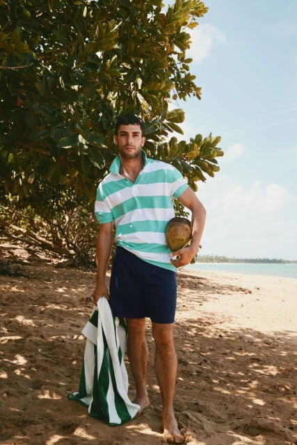 A tanned man poses on a sandy beach holding a coconut and striped towel, wearing a striped polo shirt, in sunny, outdoor setting.