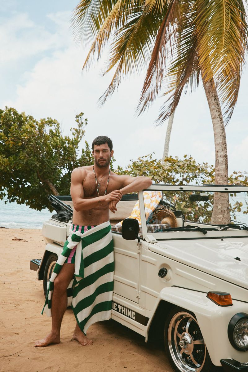 A man poses near an off-road vehicle at the beach wearing a striped towel. Tropical foliage and bright daylight create a beach scene.