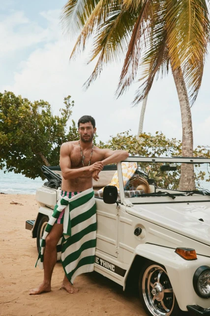 A man poses near an off-road vehicle at the beach wearing a striped towel. Tropical foliage and bright daylight create a beach scene.