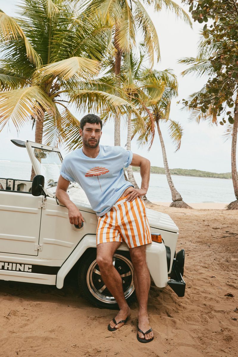 Male model poses in striped shorts and a graphic tee next to a white convertible, with palm trees and ocean in the background.