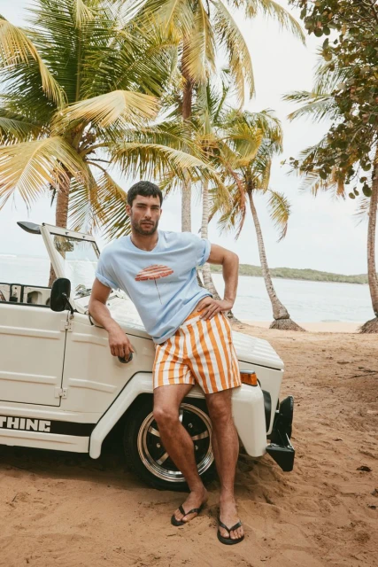 Male model poses in striped shorts and a graphic tee next to a white convertible, with palm trees and ocean in the background.