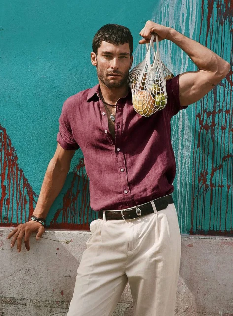 Handsome man in a casual burgundy shirt and cream pants poses with a net bag of fruit in front of an aqua graffiti wall.