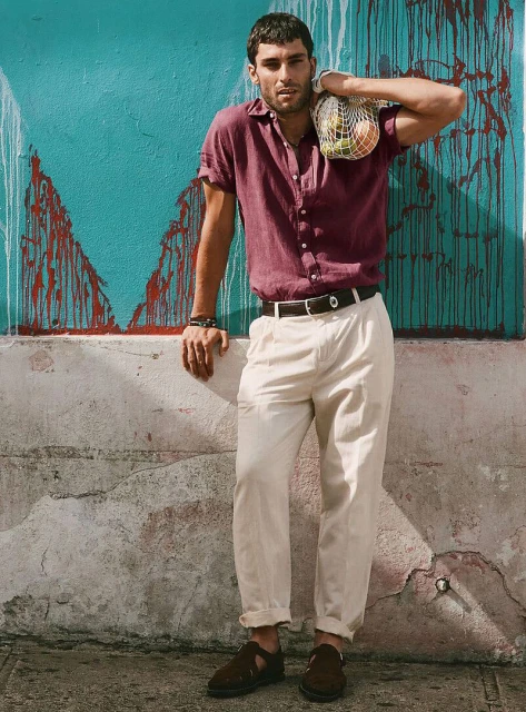 A man leans against a textured wall in casual clothing, holding a mesh bag with fruit. Shot in outdoor setting with natural light.