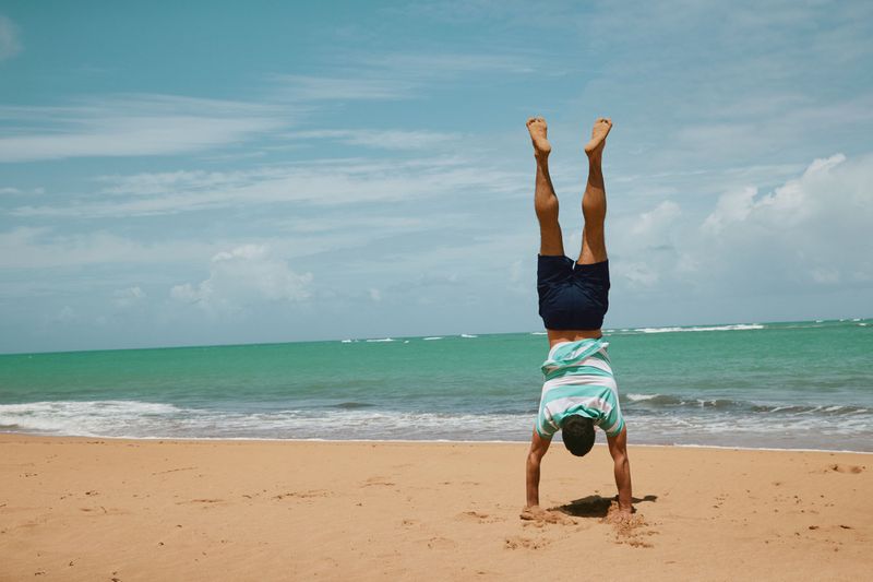 Adult does a handstand on a sandy beach by the ocean with a blue sky. The horizon is visible in the background of the outdoor scene.