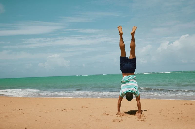 Adult does a handstand on a sandy beach by the ocean with a blue sky. The horizon is visible in the background of the outdoor scene.