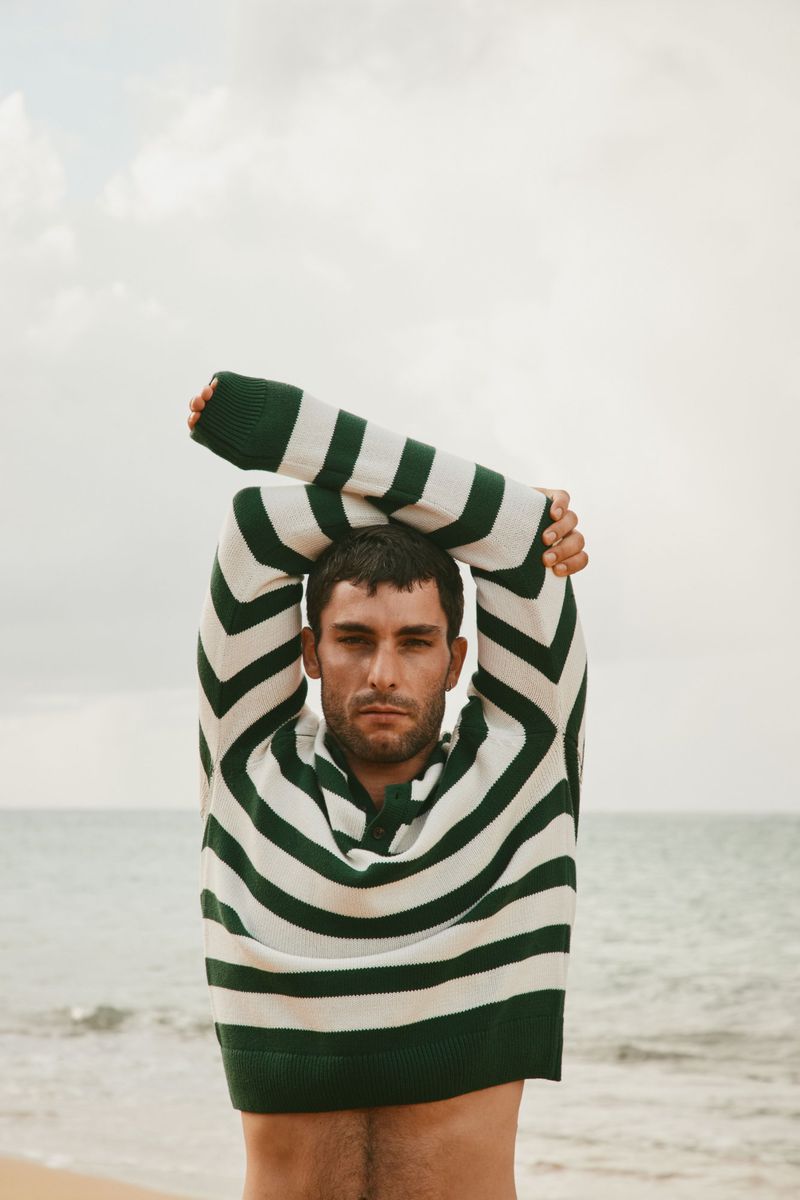 Adult man in a striped sweater stands on a sandy beach with arms overhead, the ocean visible in the background on an overcast day.