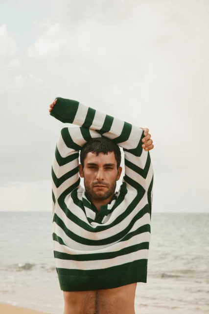 Adult man in a striped sweater stands on a sandy beach with arms overhead, the ocean visible in the background on an overcast day.
