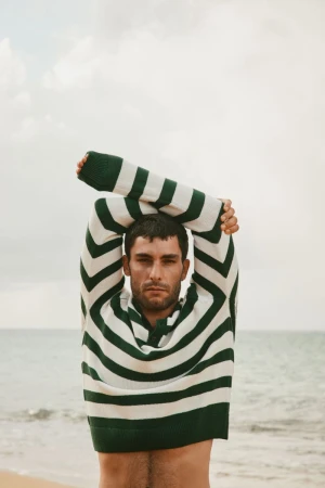 Adult man in a striped sweater stands on a sandy beach with arms overhead, the ocean visible in the background on an overcast day.