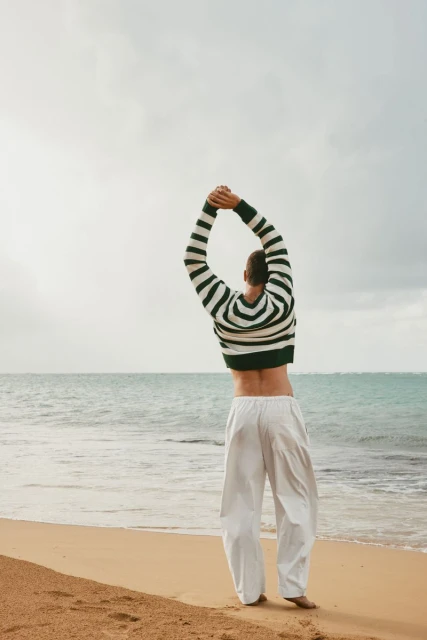 Man stretches on a beach wearing a striped sweater and white pants, with the ocean and a cloudy sky in the background.
