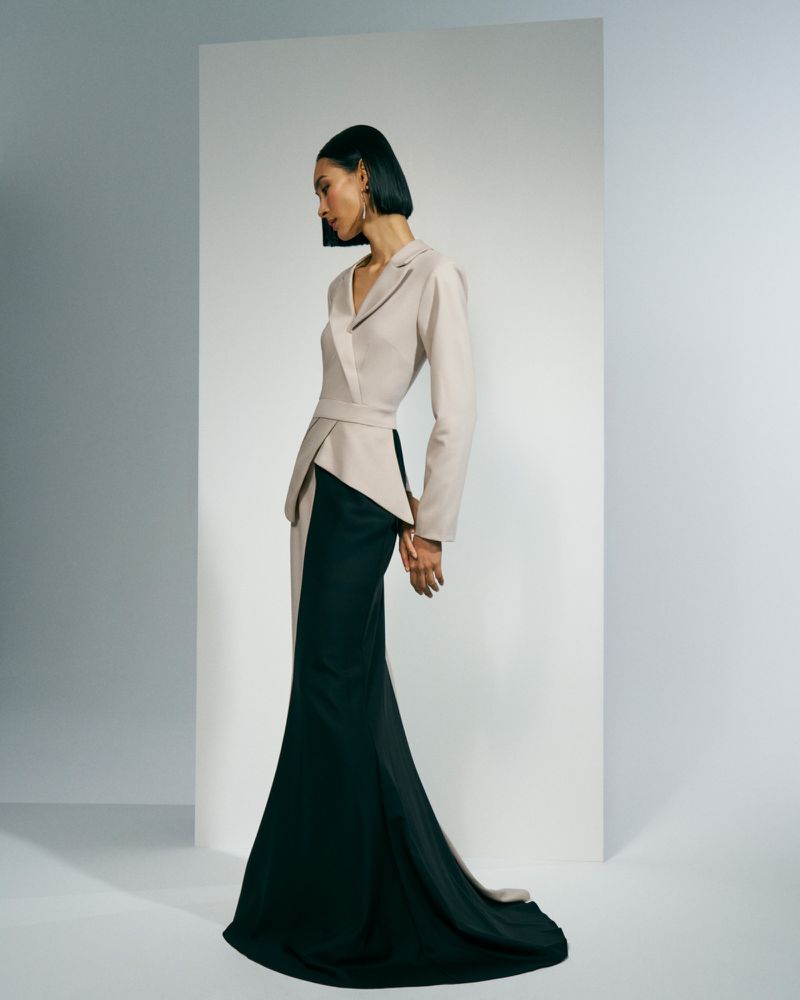 Model in a structured beige peplum blazer and sweeping black floor-length skirt posing in a minimalist studio setting.