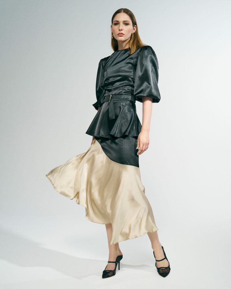 Elegant woman in a shiny black top with puffy sleeves and a beige and black skirt posing in a studio with white background.