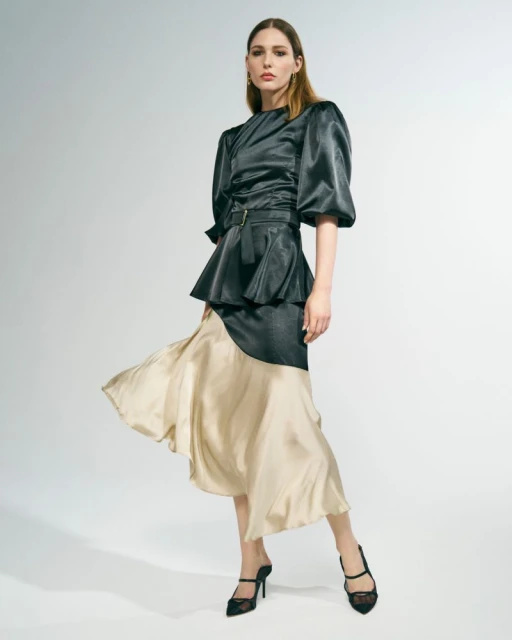 Elegant woman in a shiny black top with puffy sleeves and a beige and black skirt posing in a studio with white background.