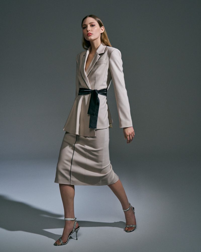 A stylish adult woman poses indoors wearing a cream-colored blazer, matching skirt, black velvet belt, and high heels in a fashion setting.