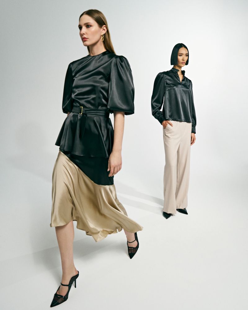 Two women model black satin tops with skirts and pants in a studio, showcasing elegant fashion against a white backdrop with sharp lighting.
