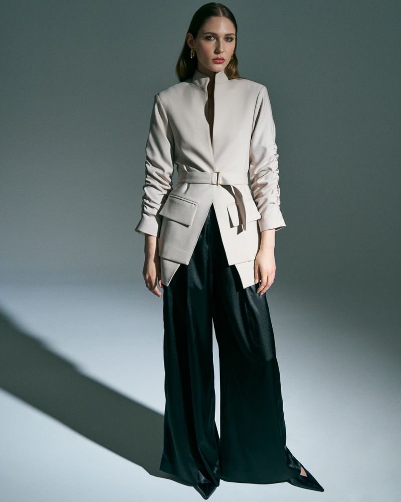 Woman in a modern beige belted jacket and flowing black trousers in a studio setting, posing elegantly with direct lighting.