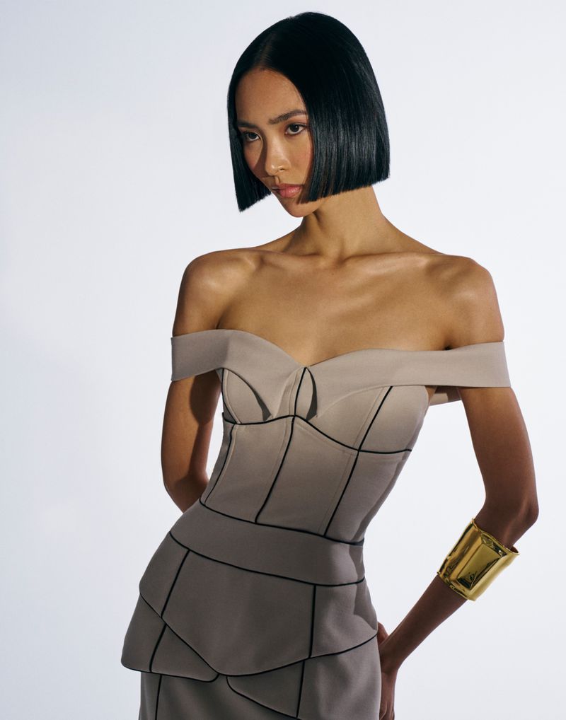 Woman with an Asian appearance poses in a studio. She is wearing an off-the-shoulder dress and a wide gold cuff bracelet on her arm.
